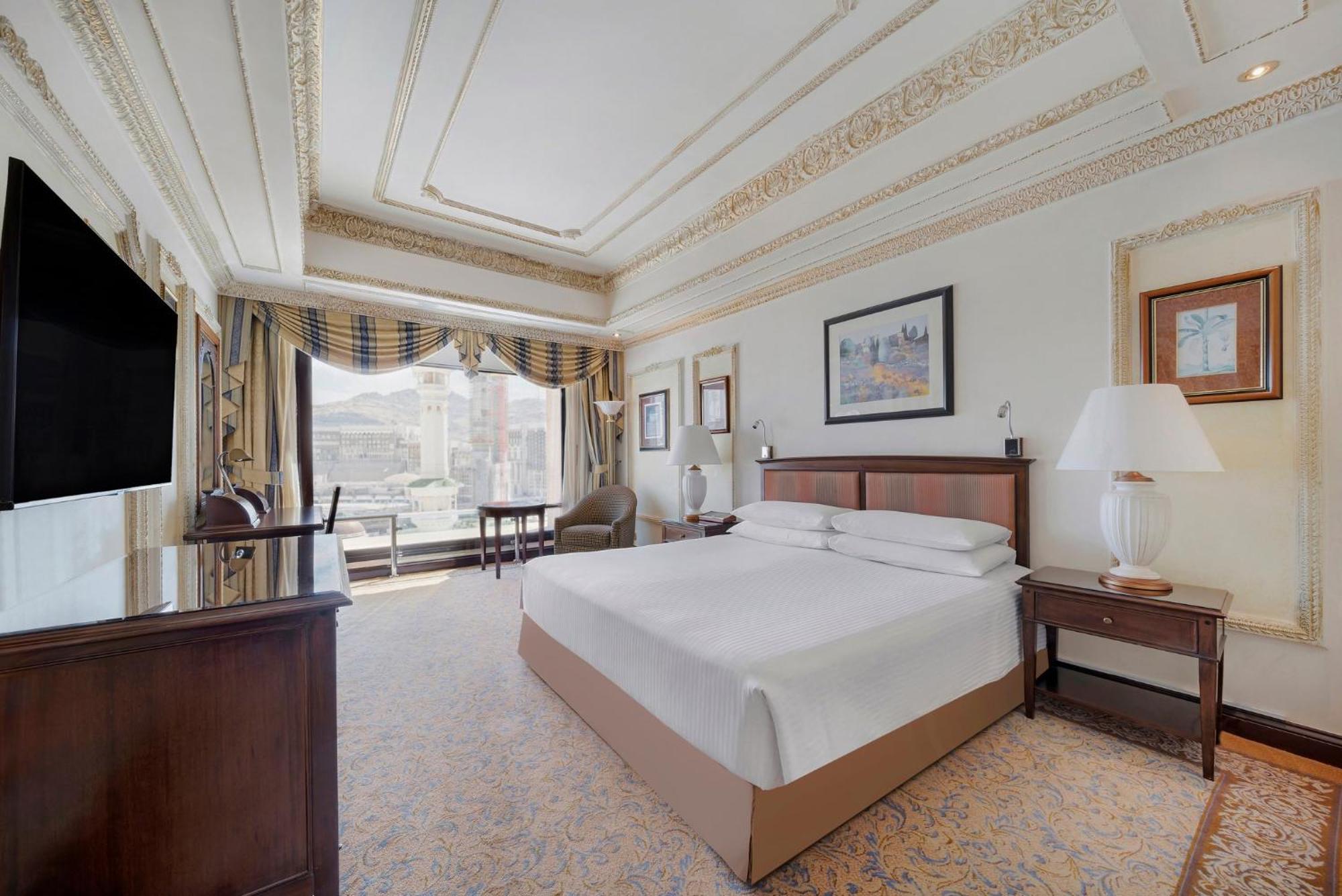 Hotel Dar Al Tawhid Intercontinental Makkah By Ihg Mekka Exterior foto The photo shows a spacious hotel room featuring a large bed with white linens. The room has elegant decor with detailed ceiling designs and soft, warm lighting. There is a wooden chest of drawers and a lamp beside the bed. A flat-screen TV is mounted