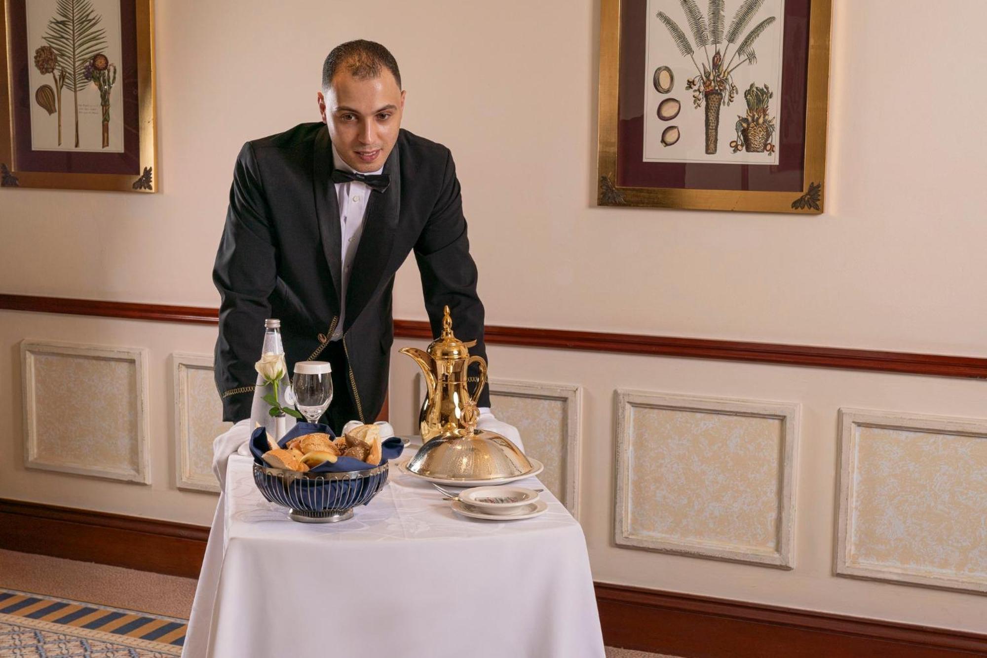 Hotel Dar Al Tawhid Intercontinental Makkah By Ihg Mekka Exterior foto The image shows a waiter dressed in formal attire, including a tuxedo. He is standing next to a white tablecloth-covered cart. On the cart, there is a covered dish, a bowl with bread rolls, a glass of water, and a bottle of champagne. The background 