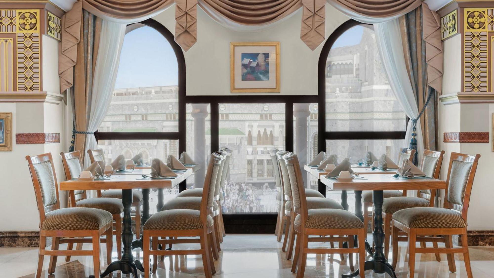 Hotel Dar Al Tawhid Intercontinental Makkah By Ihg Mekka Exterior foto The image shows a restaurant setting with several wooden tables and chairs arranged neatly. The decor features elegant curtains and drapes that frame large arched windows, allowing natural light to enter. A tasteful painting is displayed on the wall,