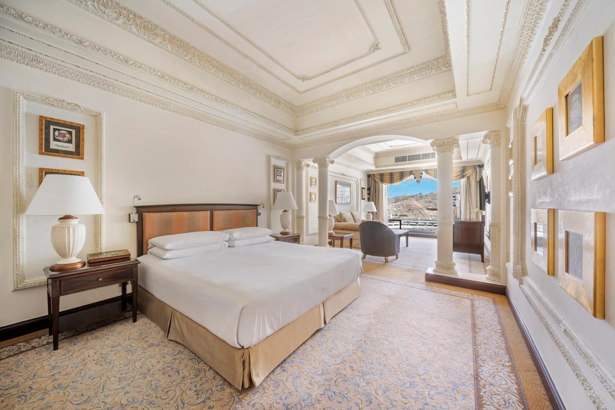 Hotel Dar Al Tawhid Intercontinental Makkah By Ihg Mekka Exterior foto The photo shows a luxurious hotel bedroom. It features a large bed with a simple, elegant design. The room includes ornate architectural details such as high ceilings and decorative moldings. There is a stylish bedside lamp on a wooden nightstand, an