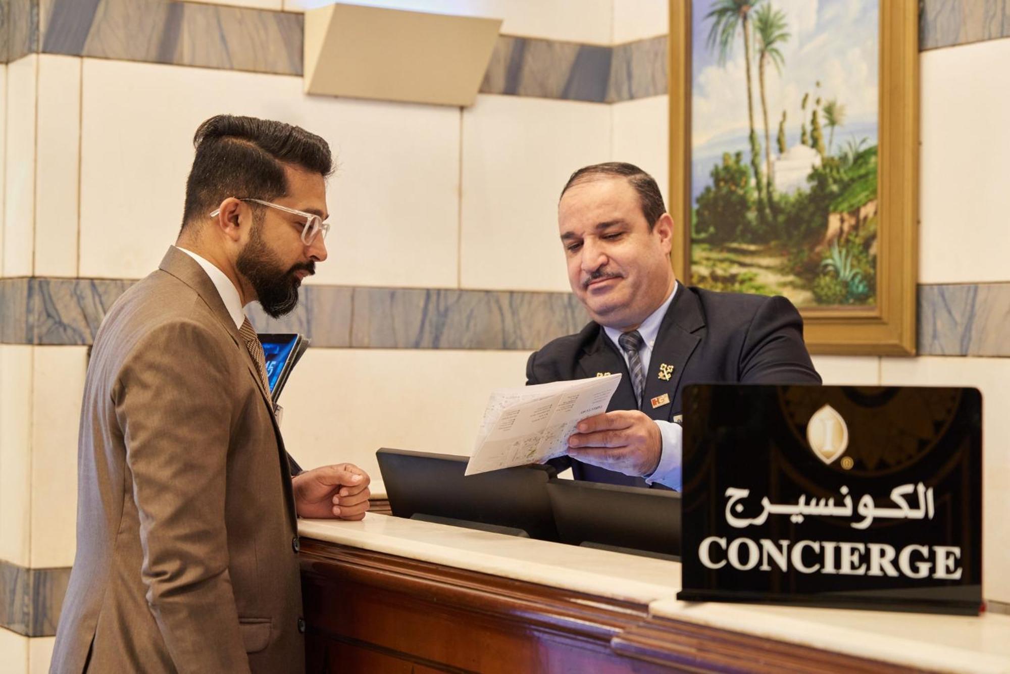 Hotel Dar Al Tawhid Intercontinental Makkah By Ihg Mekka Exterior foto The photo shows a scene at a hotel reception area. A concierge, dressed in a formal attire, is speaking with a guest who appears to be wearing a suit. The concierge is handing a document or paper to the guest, suggesting they are discussing check-in 