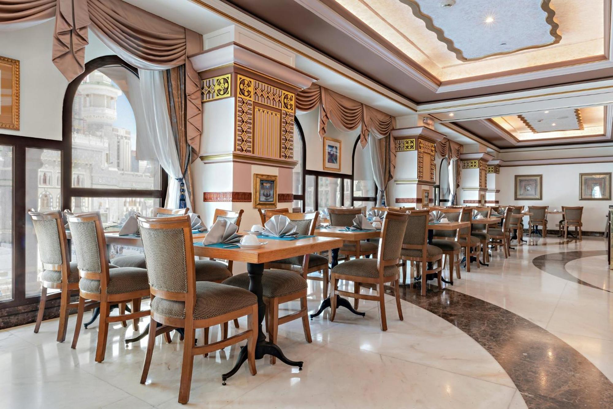 Hotel Dar Al Tawhid Intercontinental Makkah By Ihg Mekka Exterior foto The photo shows an elegantly designed restaurant interior. There are several round and rectangular wooden tables set with light blue napkins, indicating a formal dining setup. The chairs are upholstered in a neutral fabric, and the overall color sche