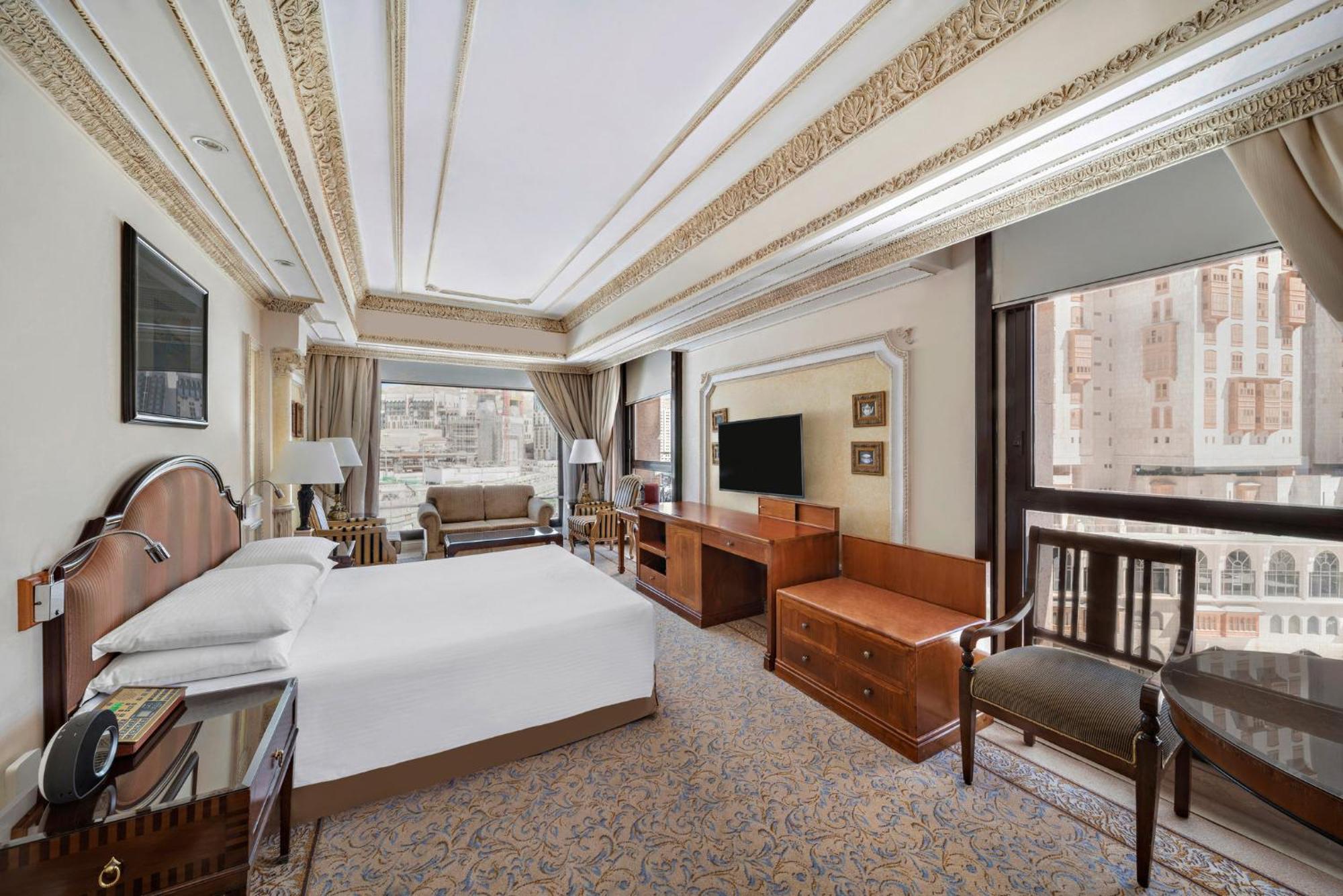 Hotel Dar Al Tawhid Intercontinental Makkah By Ihg Mekka Exterior foto The photo shows a luxurious hotel room. It features a large bed with crisp white linens, framed art on the walls, and elegant furnishings. There is a comfortable seating area with a sofa and chairs, a wooden desk, and a flat-screen TV mounted on the 