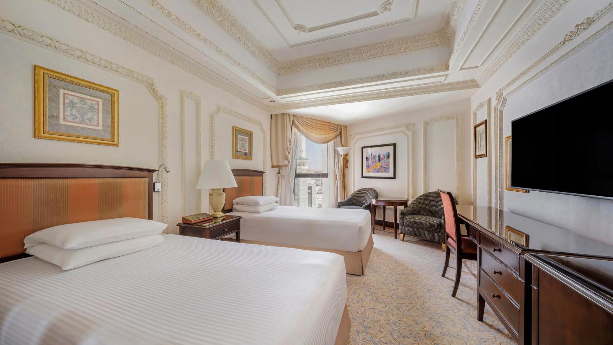 Hotel Dar Al Tawhid Intercontinental Makkah By Ihg Mekka Exterior foto The image shows an elegantly designed hotel room featuring two single beds with white linens. The walls are decorated with light colors and intricate moldings, giving a luxurious feel. There are two bedside tables with lamps beside the beds. A large 